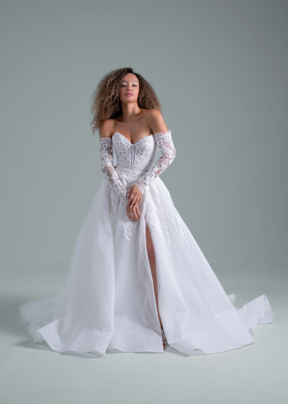 A model wearing a lace wedding dress with sleeves and a thigh-high slit.