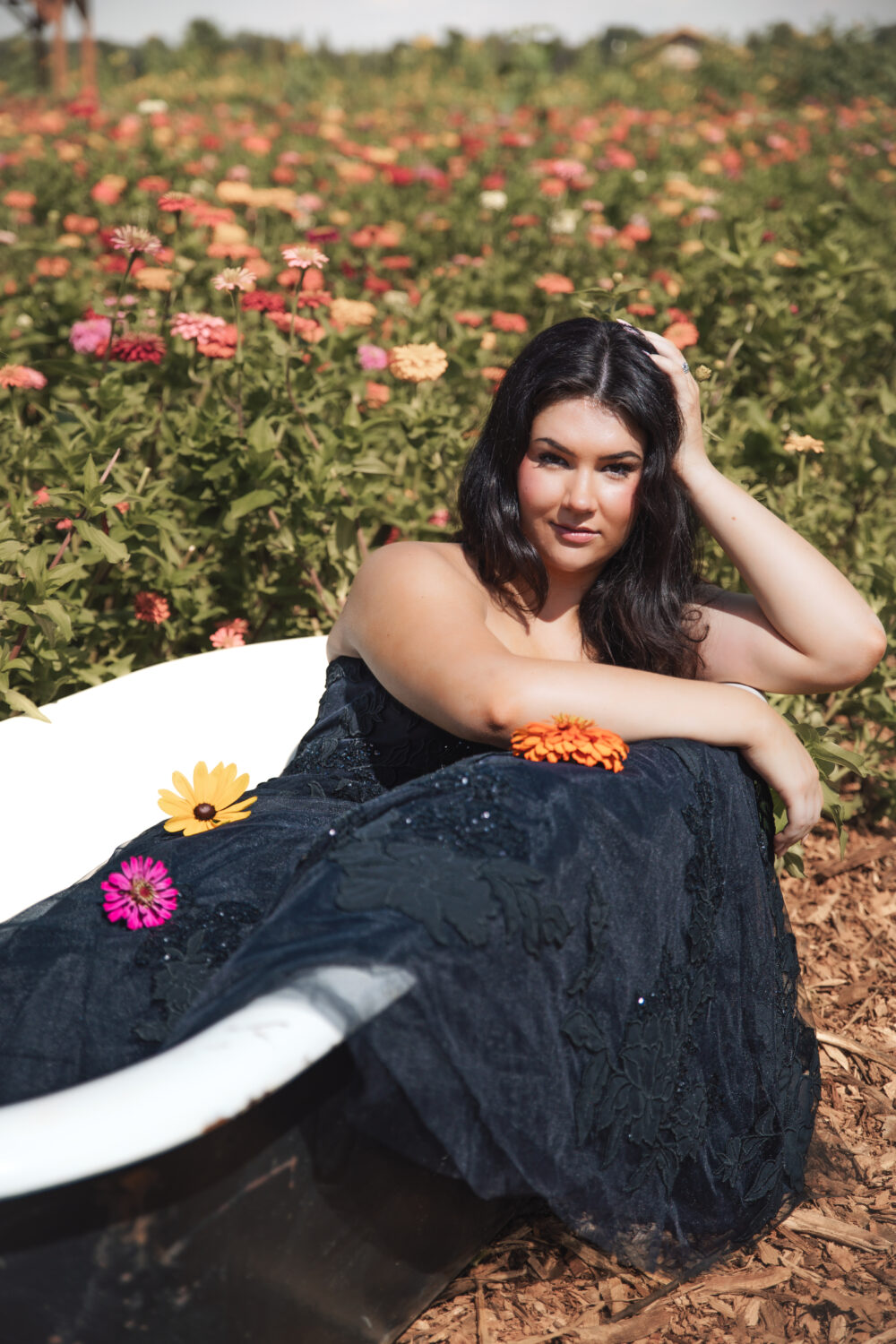 A woman in a strapless black gown lounges in a white clawfoot tub placed in a flower field, with colorful blooms resting on her dress.