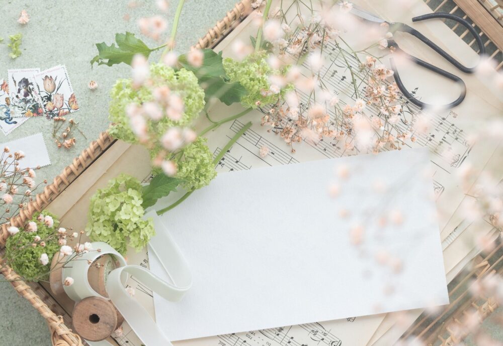 A blank white card rests on top of sheet music inside a basket, surrounded by flowers, ribbon, and a pair of scissors.