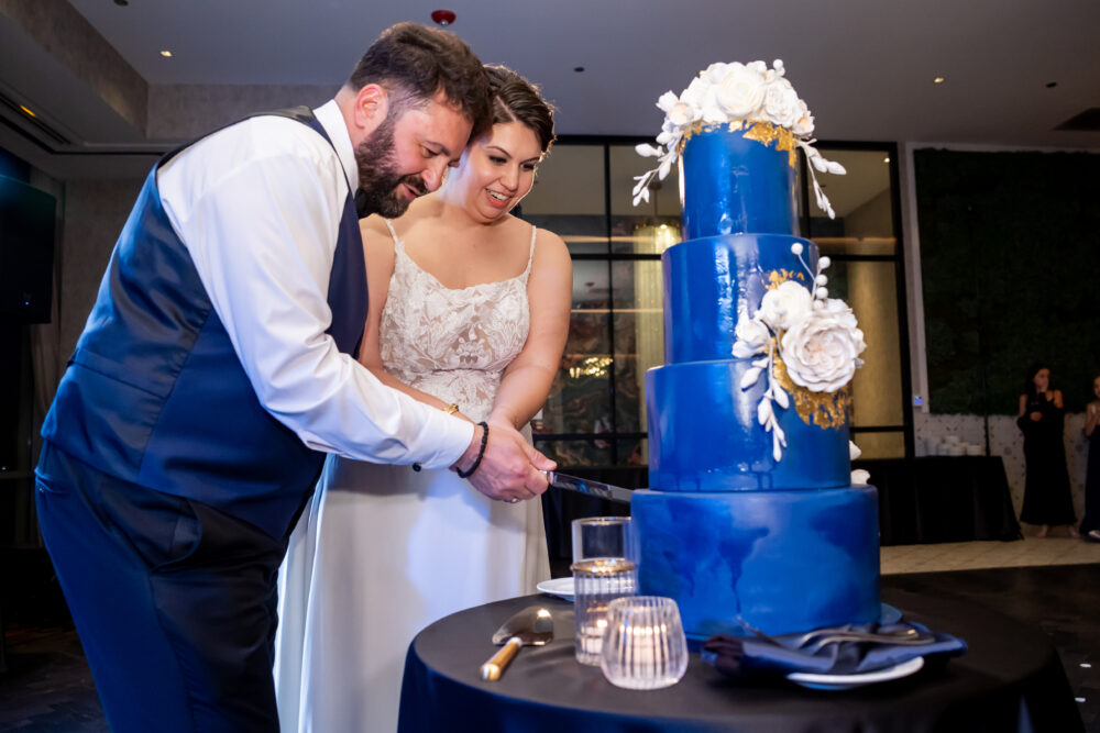 A bride and groom cut a tall, four-tier royal blue wedding cake decorated with large white sugar flowers and gold accents.