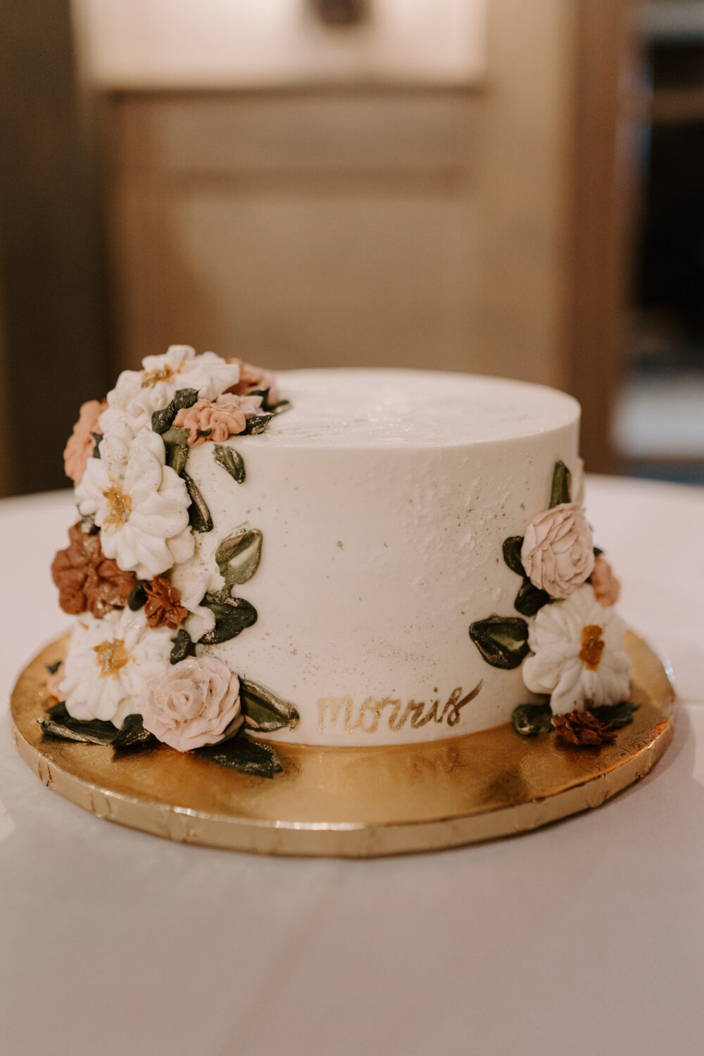 A close-up of a single-tier white wedding cake with piped floral decorations in pink, white, and green, placed on a gold cake board.