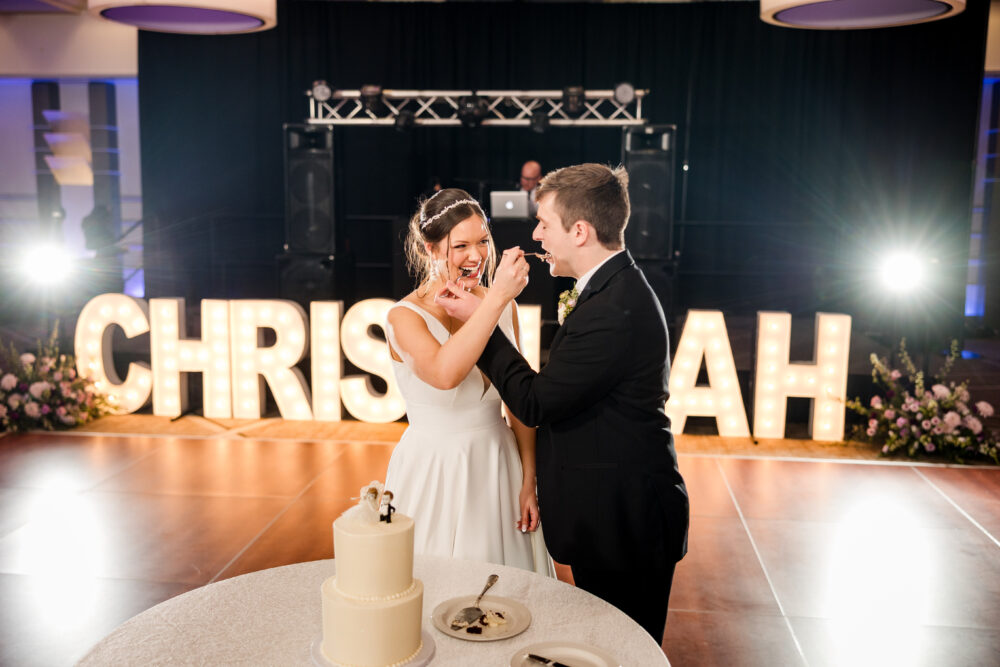 A bride and groom feed each other cake on the dance floor, standing in front of a lit-up sign.