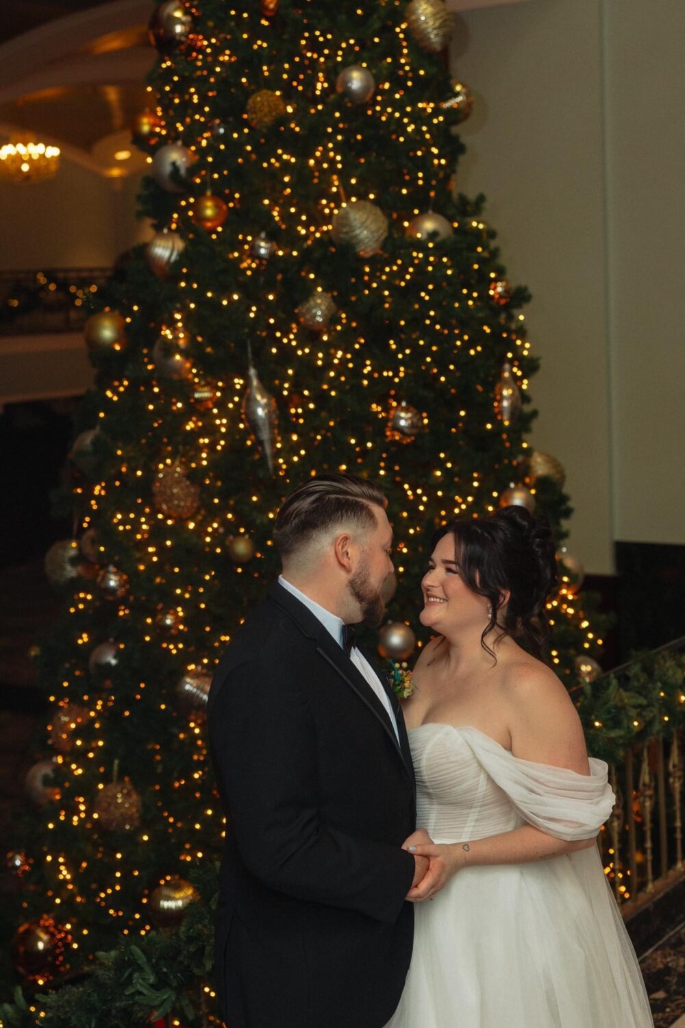 A bride and groom hold hands in front of a large Christmas tree decorated with gold ornaments and twinkling lights.