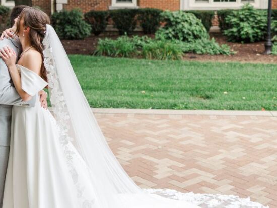 A bride and groom embrace outdoors on a brick walkway, with the bride’s long lace-trimmed train flowing behind her.