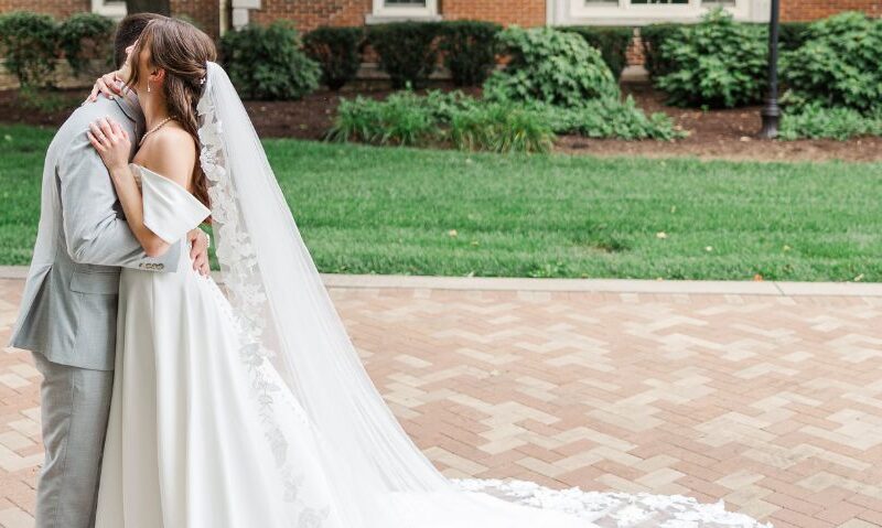 A bride and groom embrace outdoors on a brick walkway, with the bride’s long lace-trimmed train flowing behind her.
