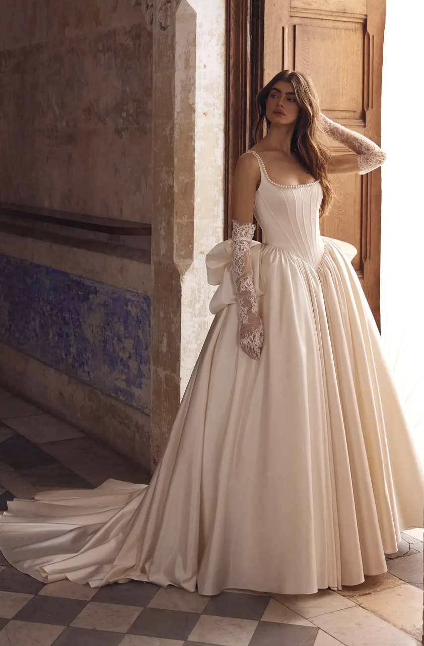 A bride stands in an elegant hall wearing a voluminous ivory gown with a pearl-trimmed neckline and lace gloves.