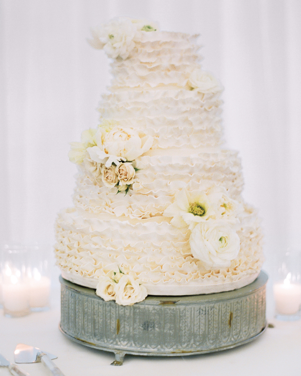 A tiered white wedding cake adorned with ivory flowers and ruffled icing, placed on a rustic metal stand.