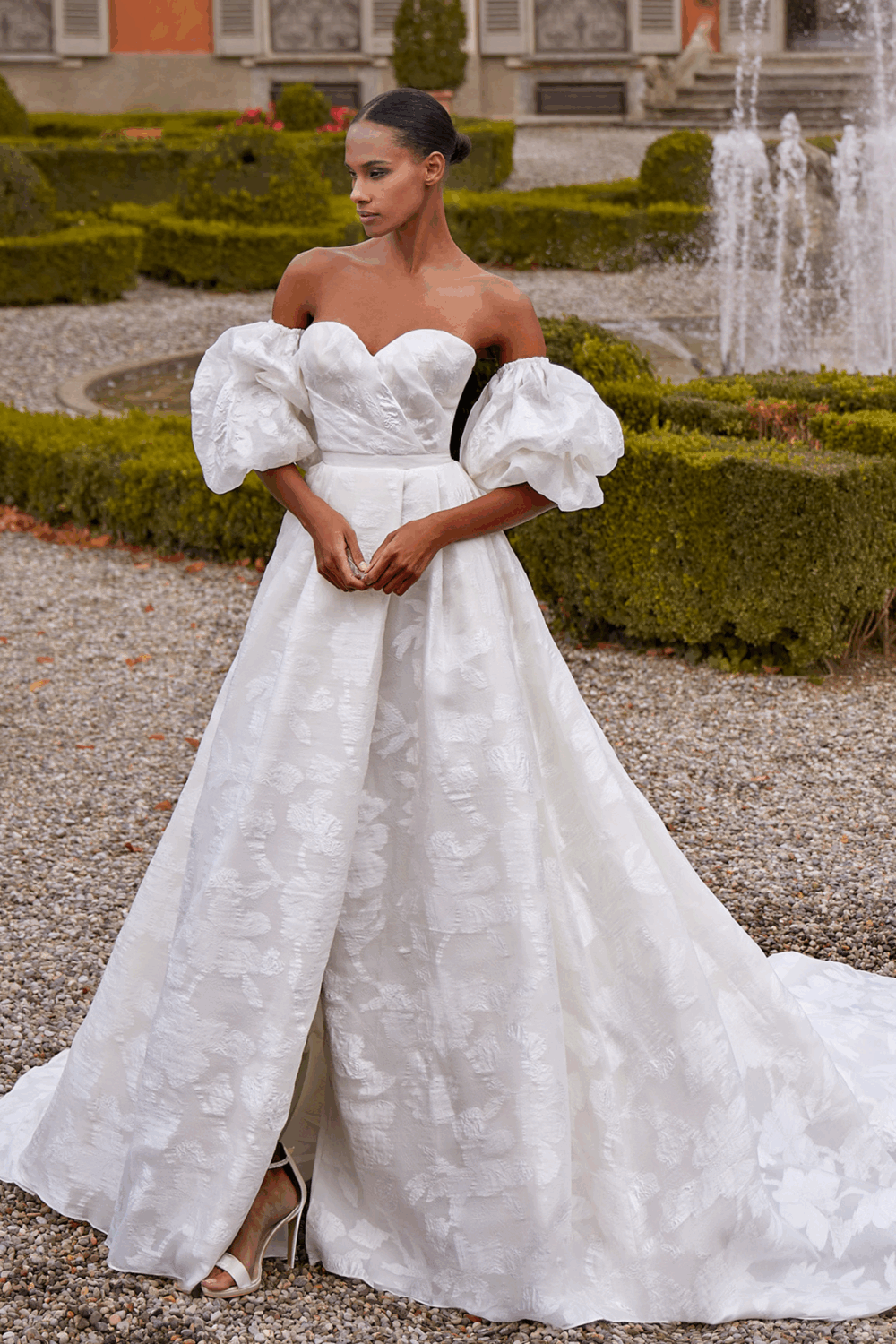 A bride stands in front of a fountain and manicured garden wearing an off-the-shoulder gown with voluminous puff sleeves.