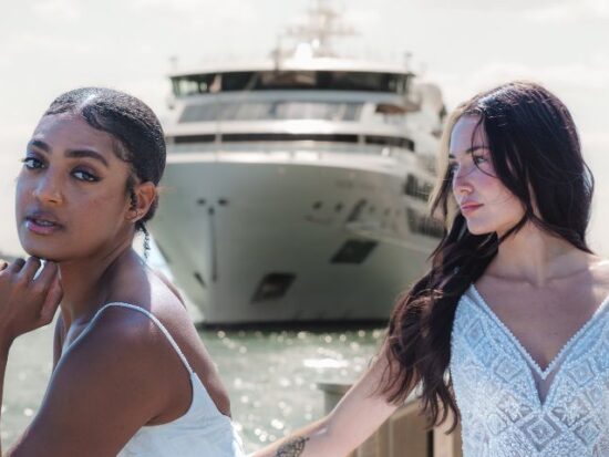 Two brides stand near the water with a large ship in the background, both wearing intricately beaded gowns.
