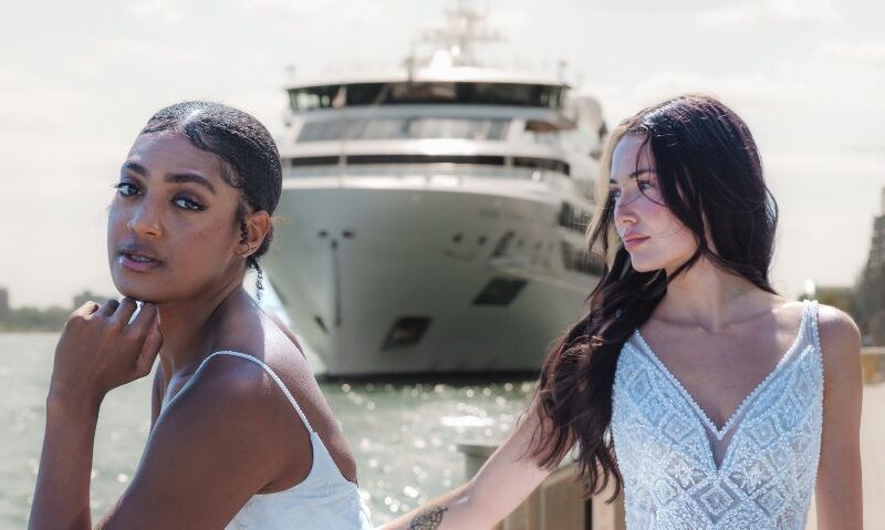 Two brides stand near the water with a large ship in the background, both wearing intricately beaded gowns.