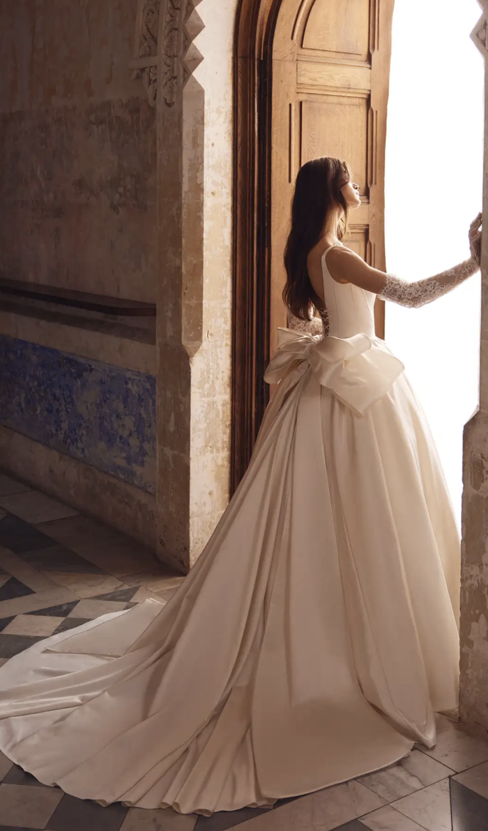 A bride stands near an arched doorway wearing a satin ballgown with a large bow and lace sleeves, softly lit by daylight.
