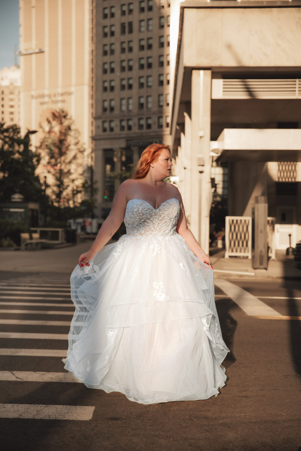 A bride with red hair poses in a strapless lace ballgown on a city street surrounded by tall buildings in warm light.