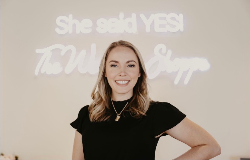 A woman wearing a black shirt stands smiling in front of a neon sign that reads "She said YES!," inside The Wedding Shoppe Boutique.