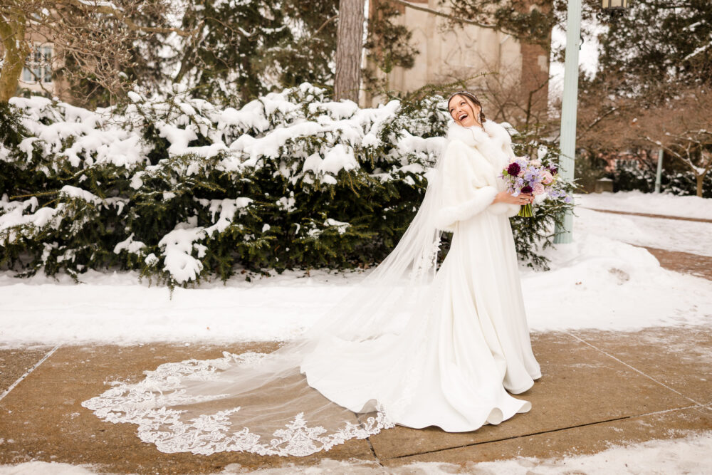A bride laughs outdoors in the snow wearing a long white gown, fur shawl, and veil while holding a pastel bouquet.