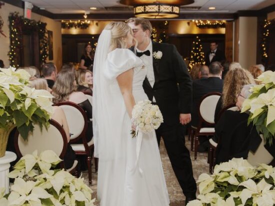A bride and groom kiss in an indoor wedding ceremony decorated with gold vases of white poinsettias and soft lighting.
