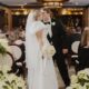 A bride and groom kiss in an indoor wedding ceremony decorated with gold vases of white poinsettias and soft lighting.