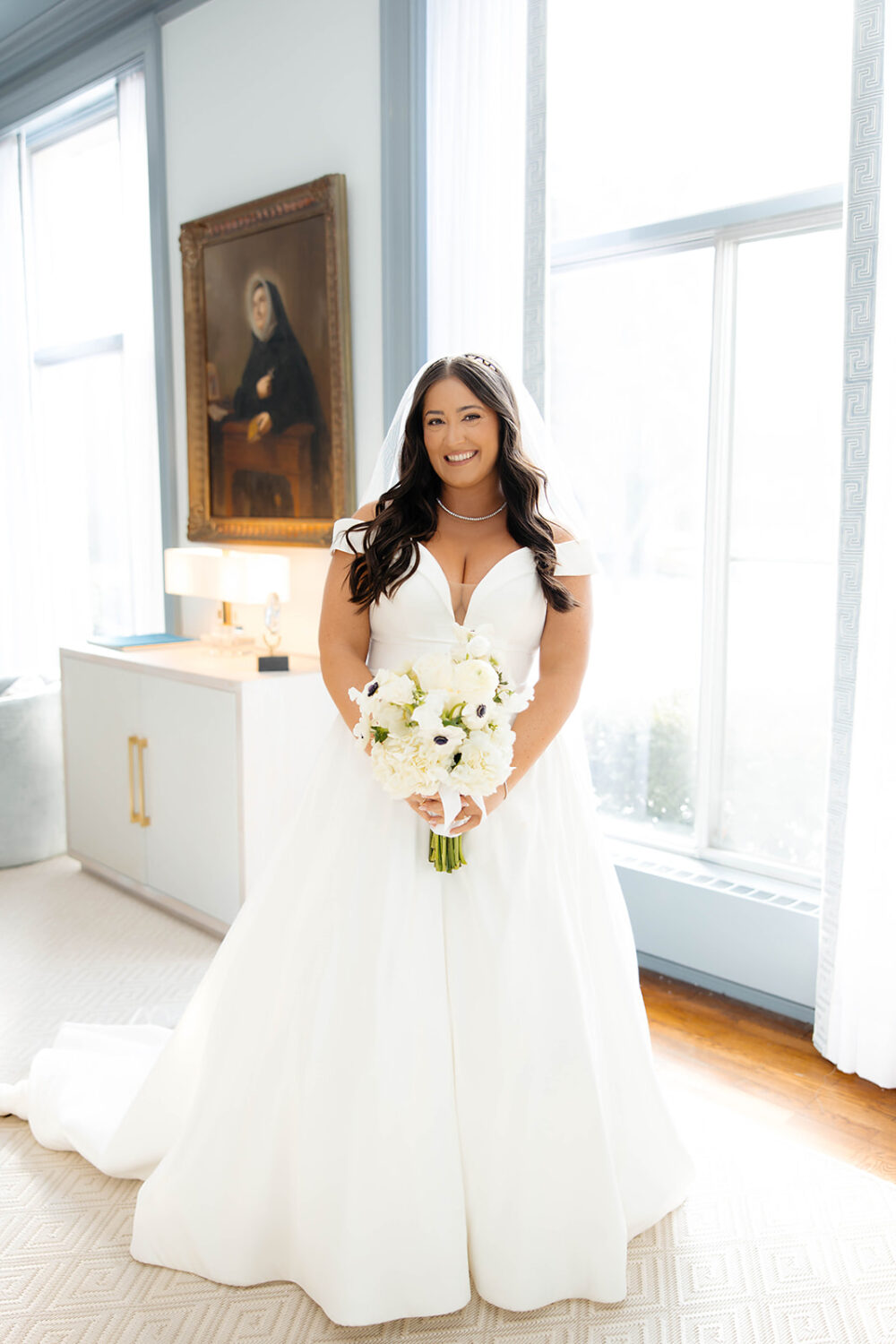 A bride stands in front of large windows holding a white bouquet, wearing an off-the-shoulder wedding dress and a veil.