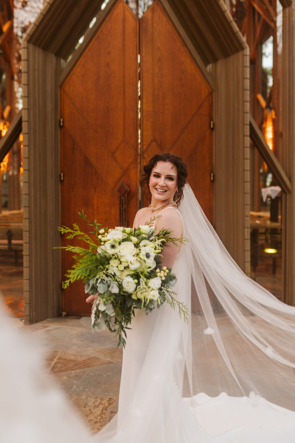 A bride stands outside a chapel with large wooden doors, holding a cascading bouquet of white flowers and greenery.