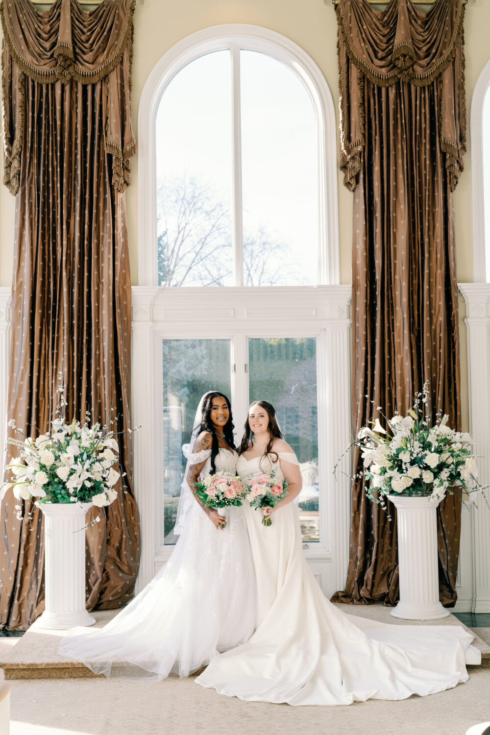 Two brides stand together in front of tall arched windows holding pink and white bouquets in an elegant indoor venue.