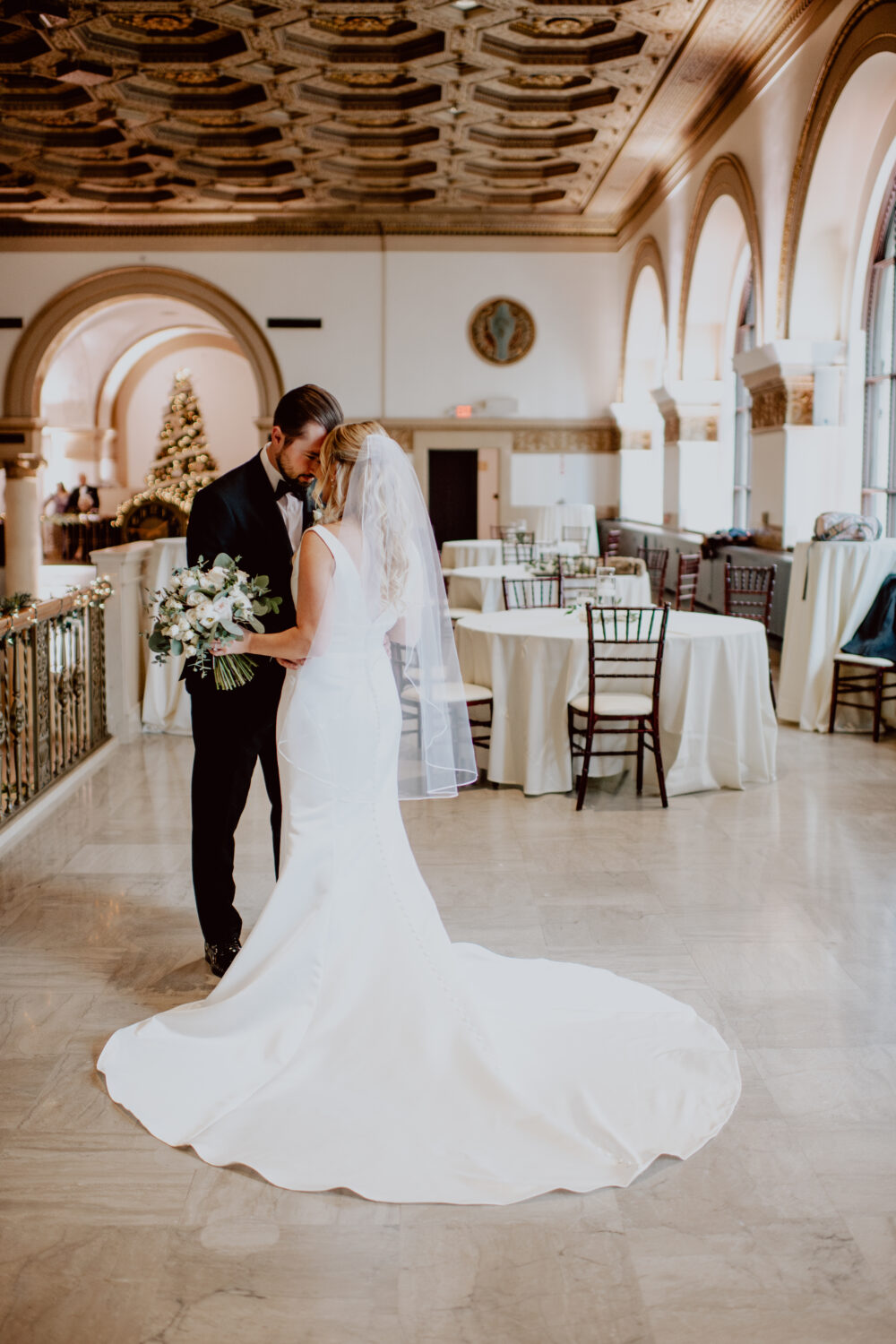 A bride and groom stand in an elegant hall with arched windows, exchanging a quiet moment with a decorated Christmas tree in the background.