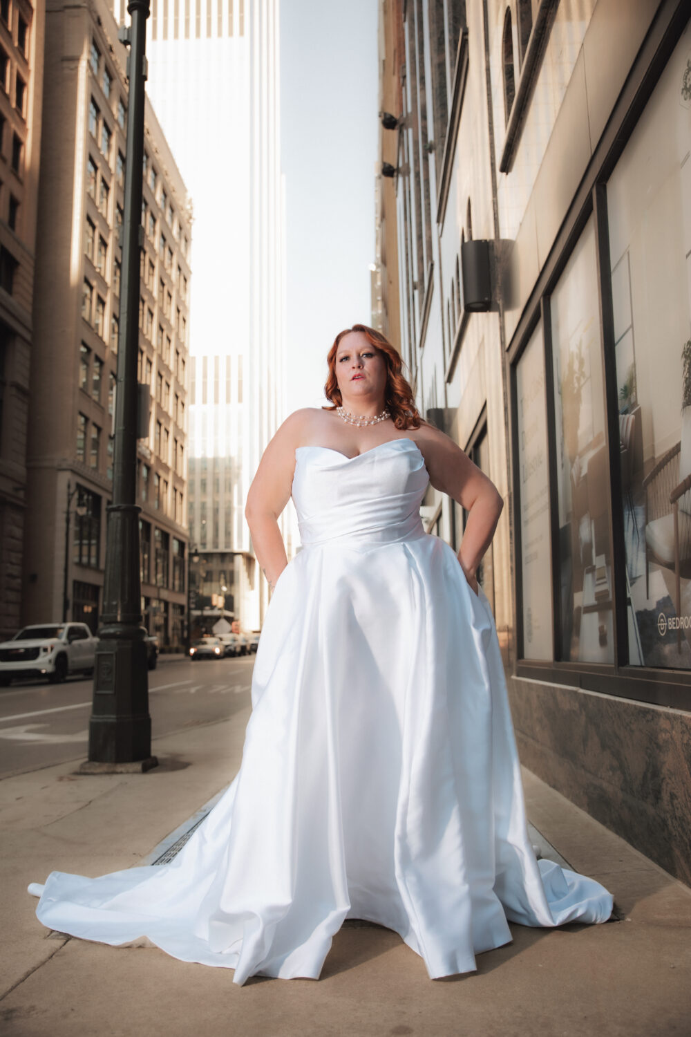 A woman in a strapless white ballgown stands on a city sidewalk between tall buildings, looking directly at the camera with cars and skyscrapers behind her.