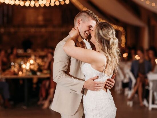 A bride and groom share a close embrace during their first dance in a warmly lit reception hall, with string lights glowing behind them and guests seated in the background.