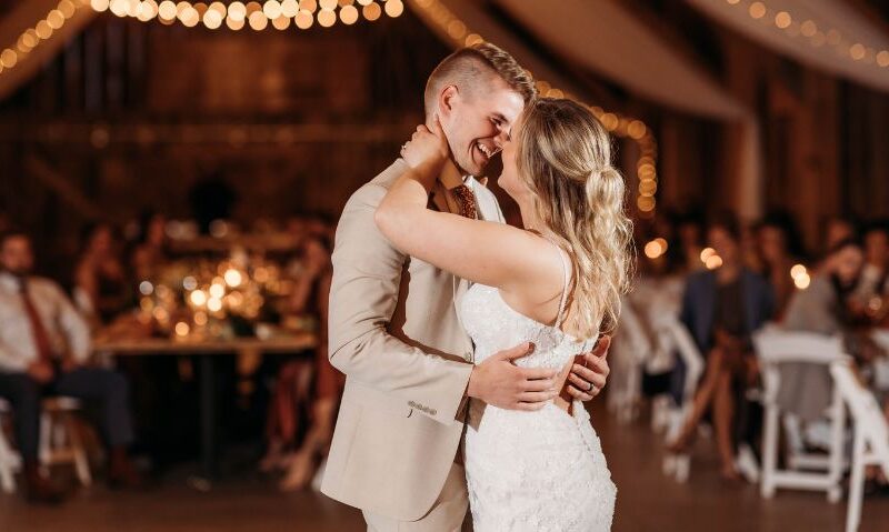 A bride and groom share a close embrace during their first dance in a warmly lit reception hall, with string lights glowing behind them and guests seated in the background.