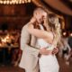 A bride and groom share a close embrace during their first dance in a warmly lit reception hall, with string lights glowing behind them and guests seated in the background.