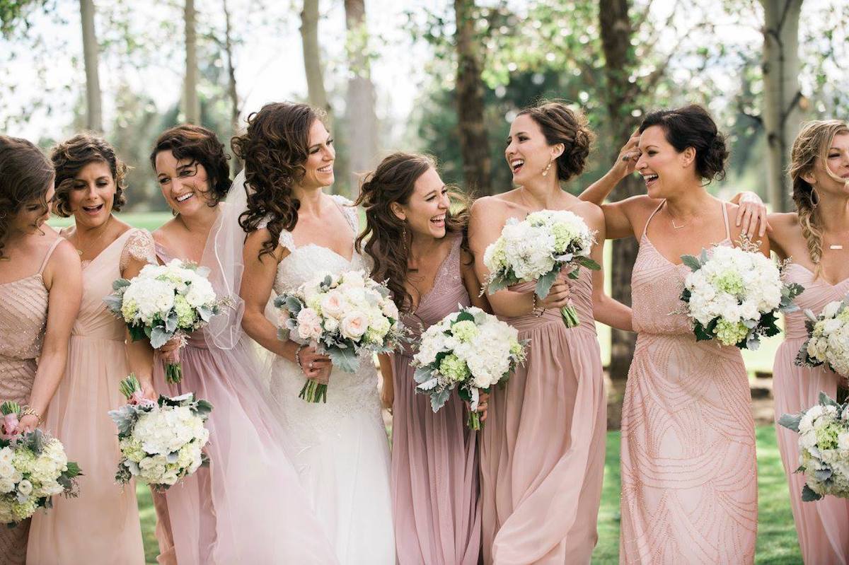 Bridesmaids in blush dresses laughing and holding white floral bouquets.