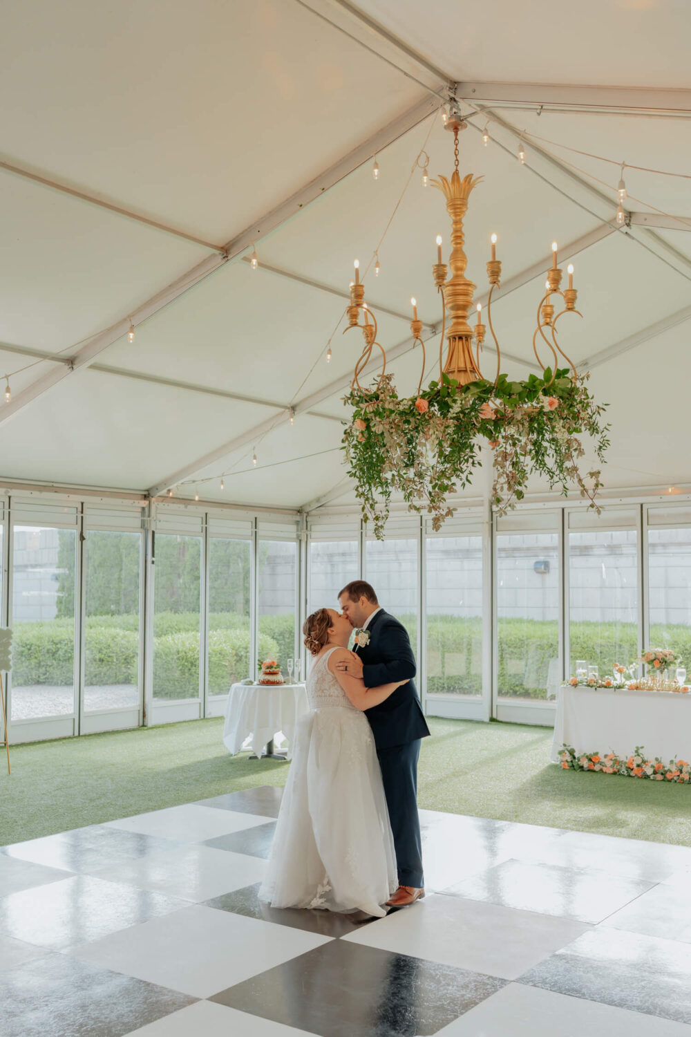 A bride and groom share a first dance on a black-and-white checkered dance floor inside a bright venue with a large golden chandelier.