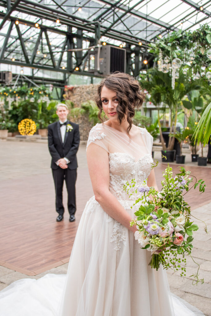 Bride smiling with bouquet on outdoor pathway near greenhouse.