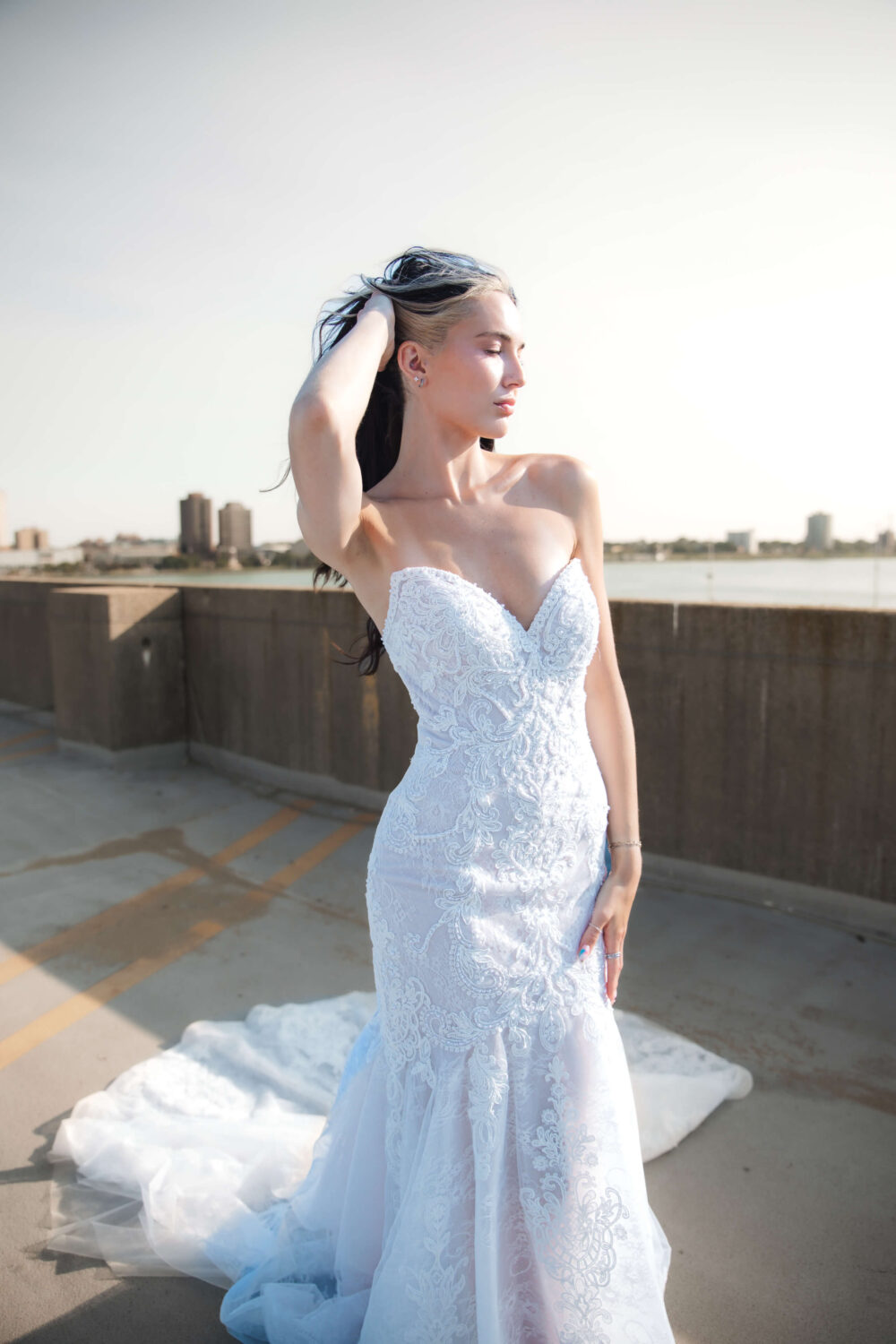 A woman in a strapless, lace-embroidered white wedding gown stands on an outdoor parking area, gently touching her hair as the sunlight illuminates her face and dress.