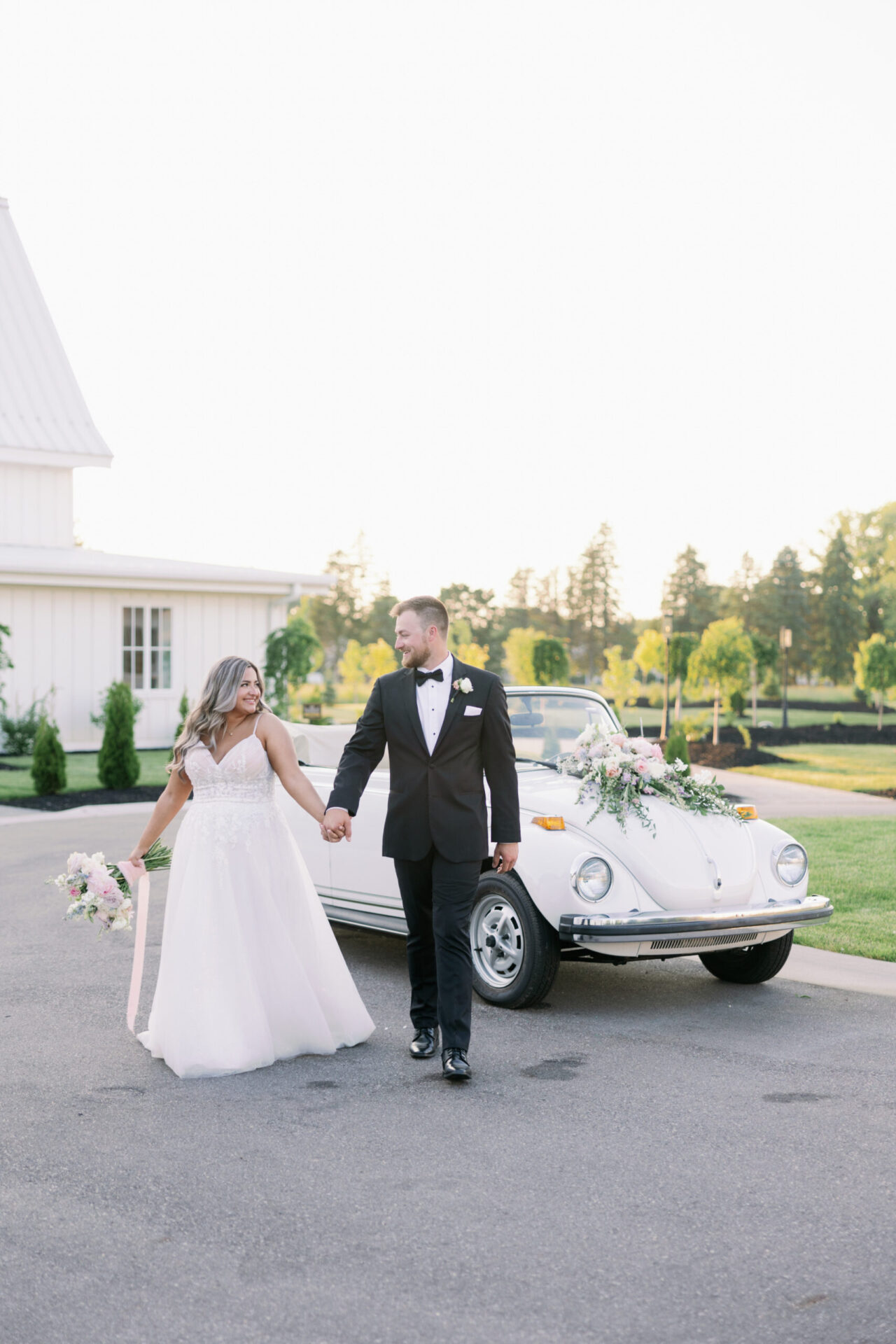 Bride and groom walking beside a vintage white car outside a house.