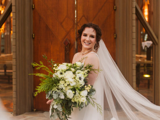Bride standing in front of wooden chapel doors with her veil blowing behind her.