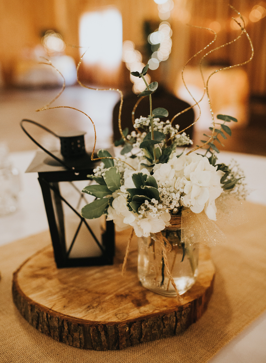Lantern and floral centerpiece decor on a wooden slice at a rustic wedding reception table.