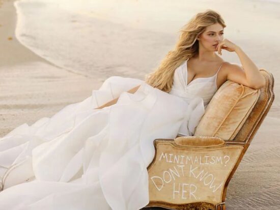 A bride lounges on a vintage chair at the beach, wearing a flowing white gown, with waves and sand in the background.