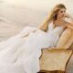 A bride lounges on a vintage chair at the beach, wearing a flowing white gown, with waves and sand in the background.