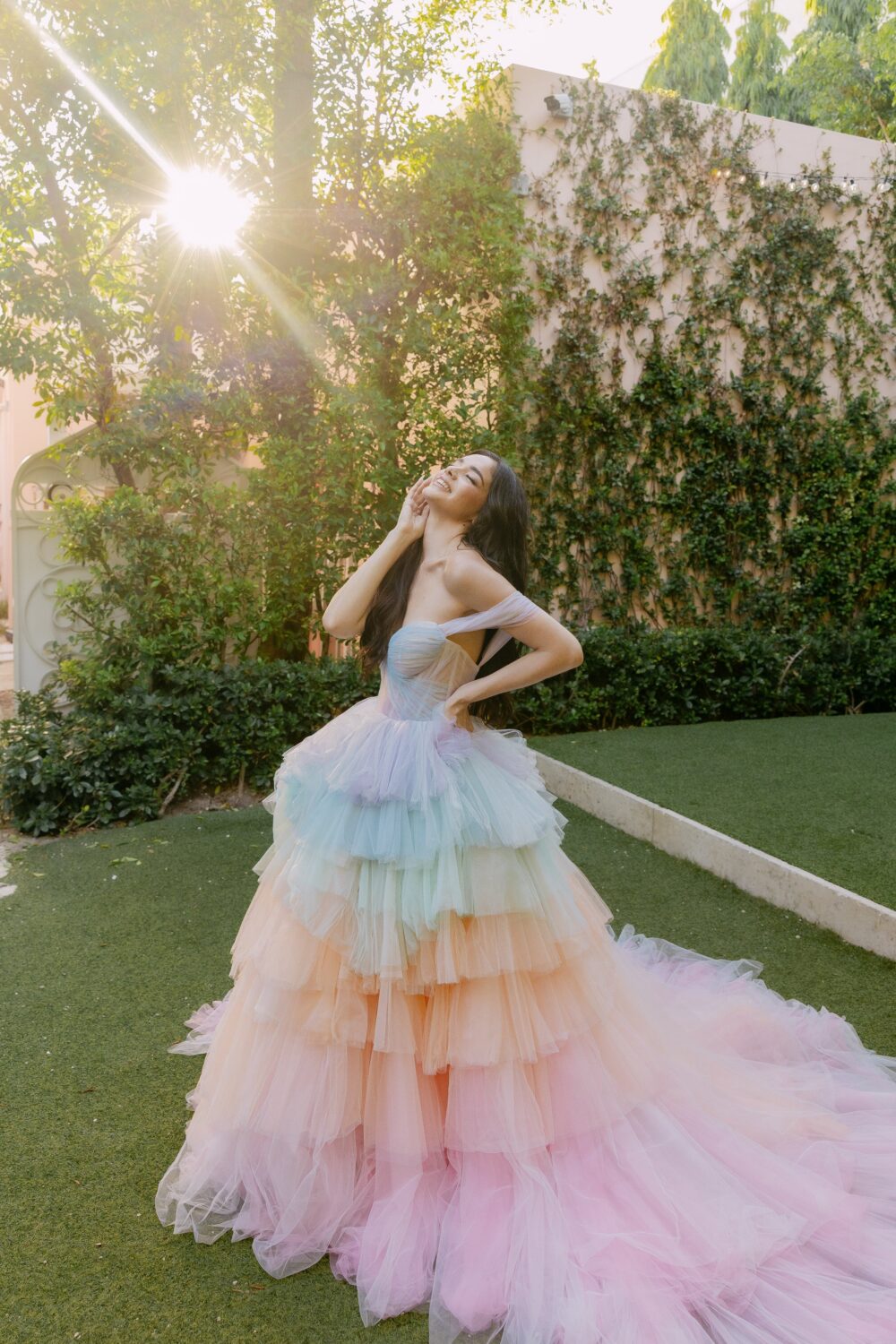 A woman in a rainbow tiered tulle ball gown stands on a sunlit lawn, smiling and tilting her head back while posing with one hand on her hip.