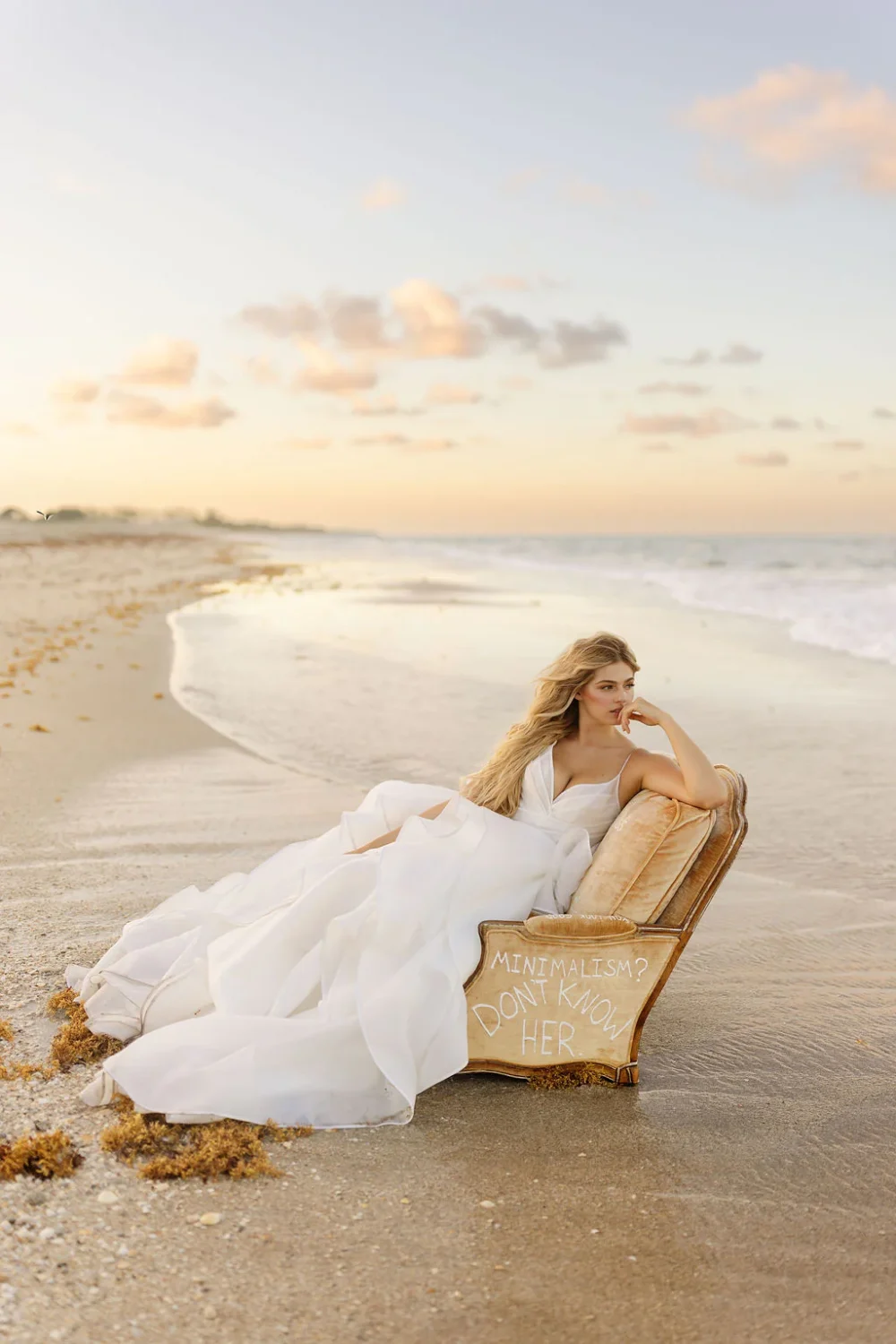 A bride in a voluminous ruffled white gown reclines on a chair at the shoreline during sunset, with soft waves and a pastel sky in the background.
