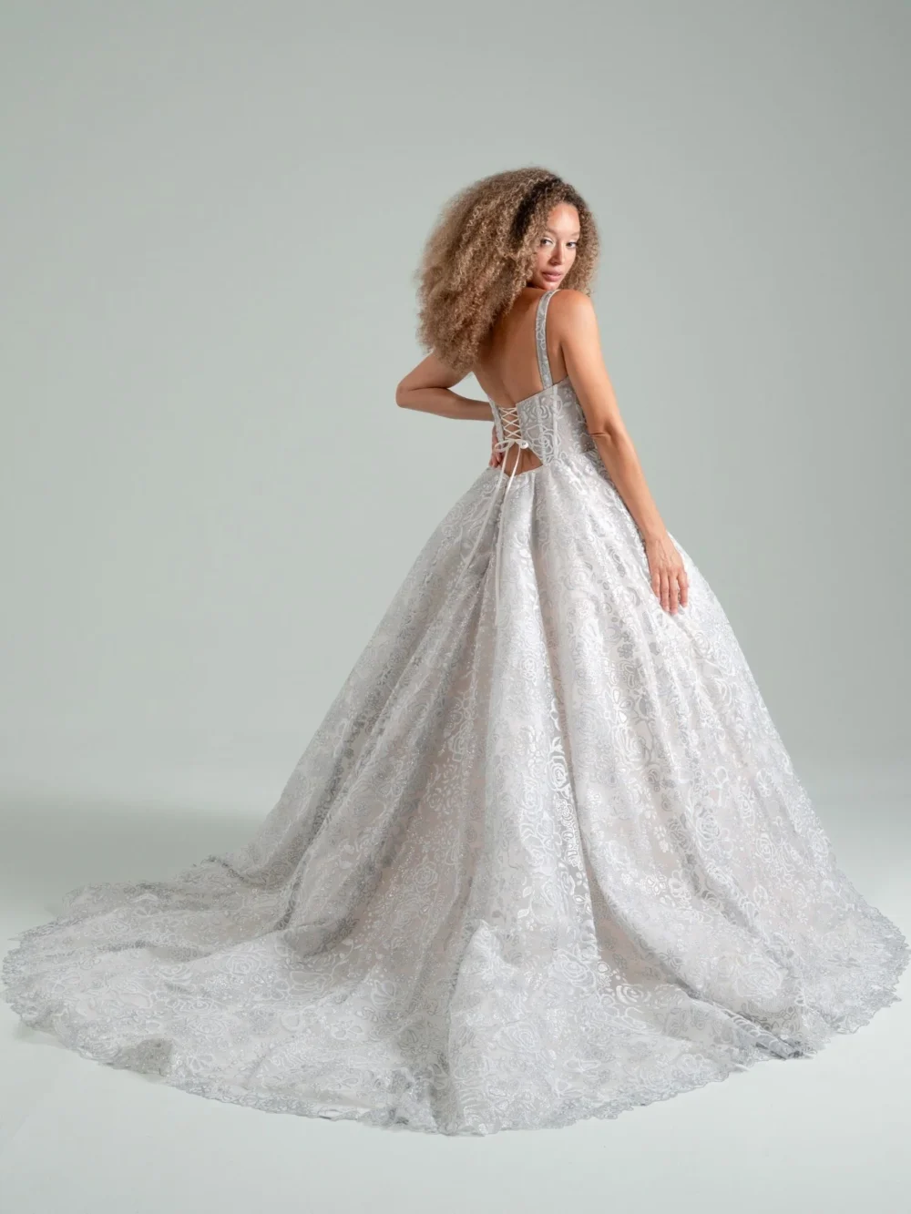A model in a silver lace ball gown with a long train glances back over her shoulder, showing the corset-style lacing on the back of the dress.