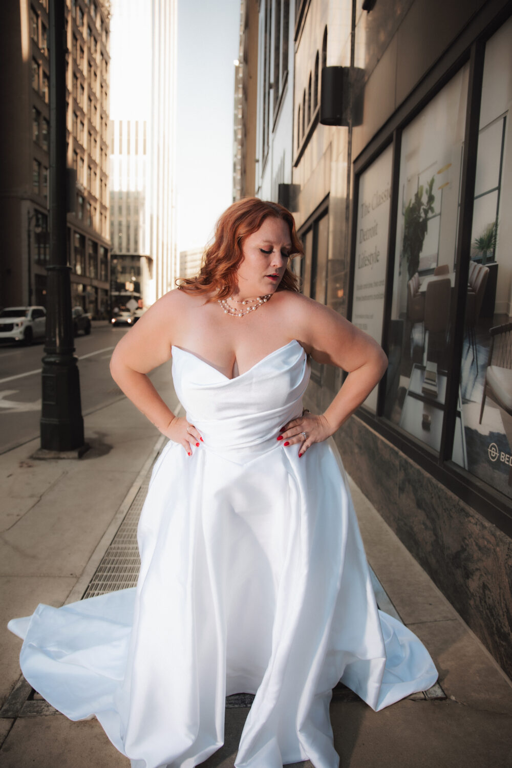 A woman in a strapless white wedding dress looks down while standing on a city sidewalk, with her hands on her waist and tall buildings rising behind her.