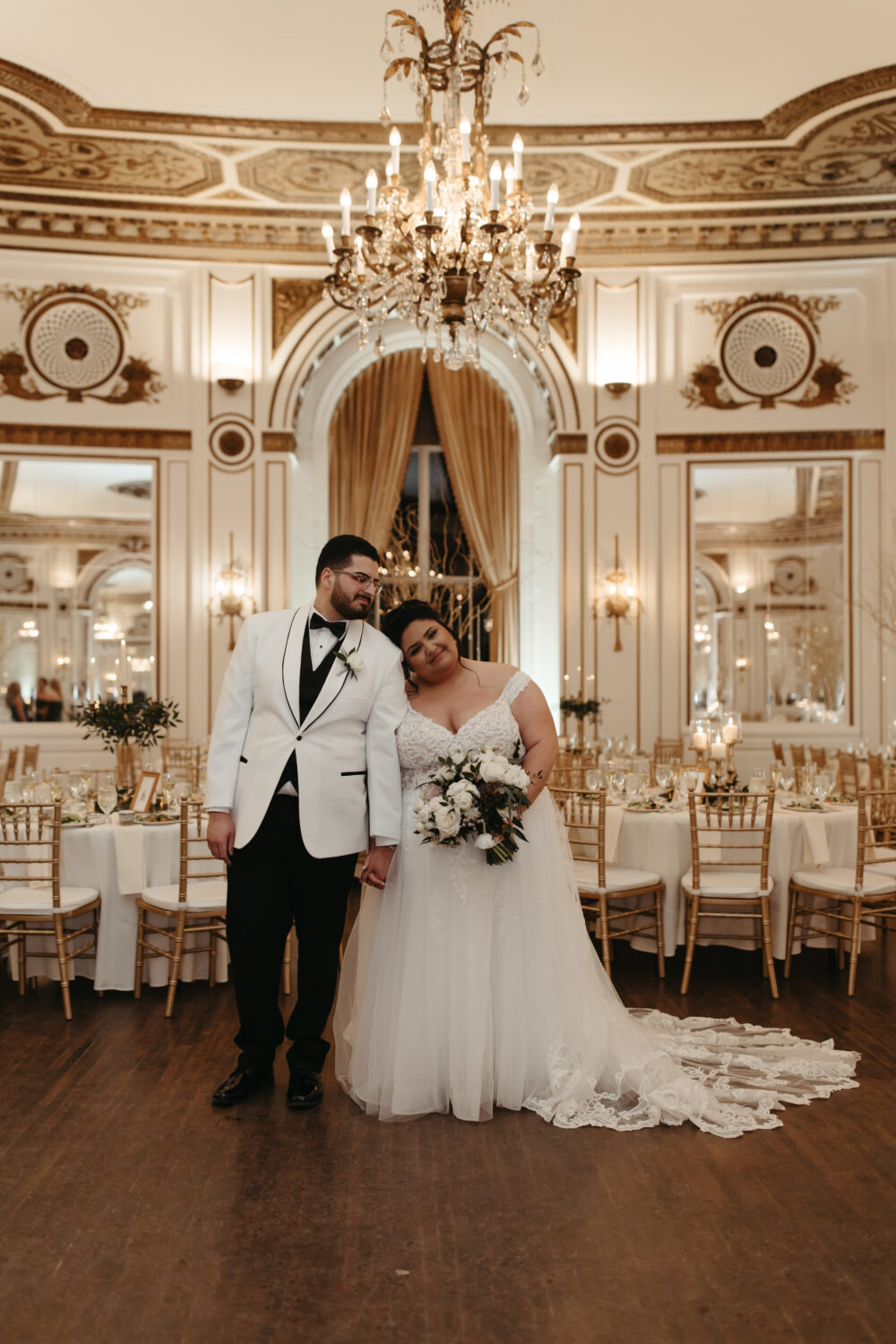 A bride and groom stand together in an elegant ballroom with chandeliers and gold decor, the bride wearing a lace wedding gown and holding a bouquet while leaning her head on the groom’s shoulder.