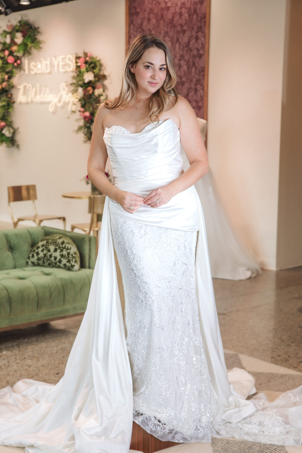 A bride wearing a strapless satin and lace gown with a long draped train poses in a bridal boutique with floral décor behind her.