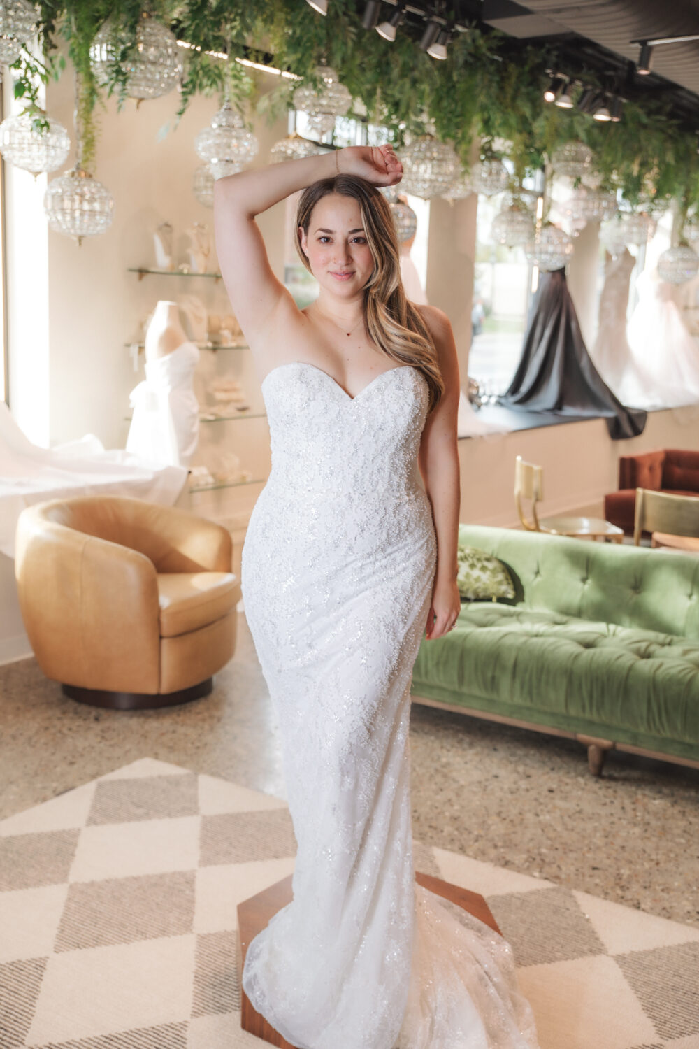 A bride in a strapless, fitted, sparkly lace wedding gown poses on a wooden pedestal in a decorated bridal boutique.