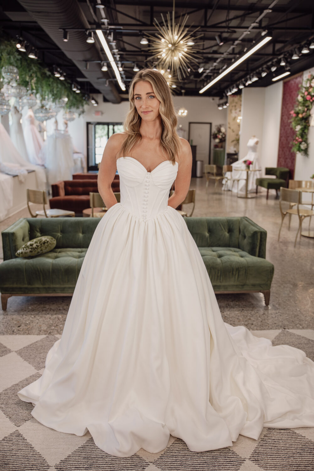 A bride wearing a strapless satin ball gown with a structured bodice stands in the middle of a bridal showroom.