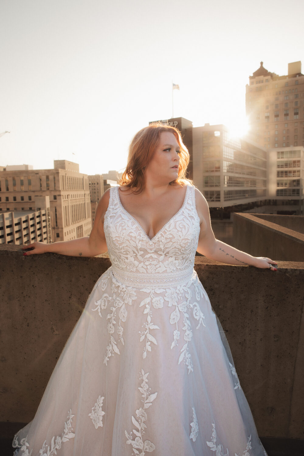 A woman in a lace A-line wedding gown stands on a rooftop at sunset, holding the ledge as the sun shines behind nearby city buildings.