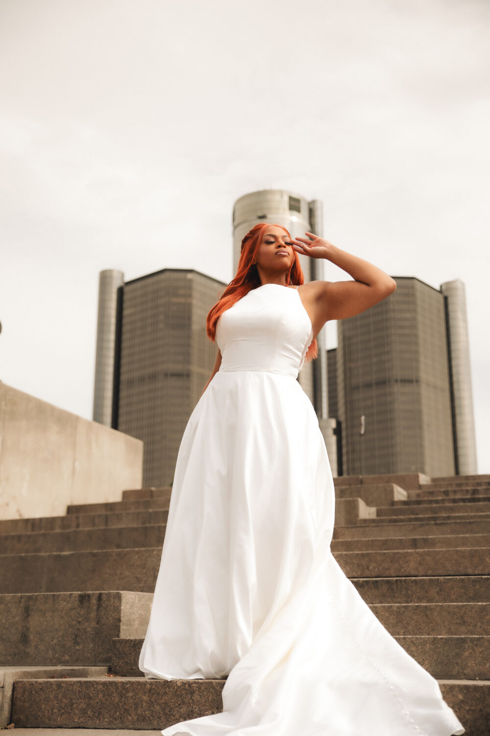 A woman with long red hair stands on outdoor steps wearing a sleeveless white wedding gown, with tall modern buildings rising behind her.