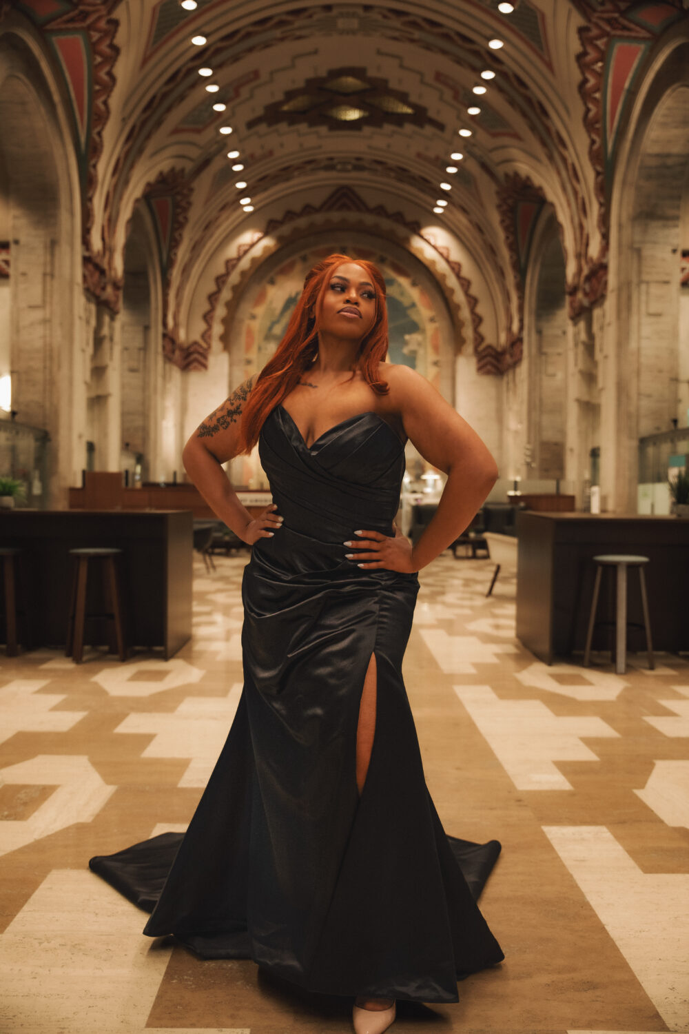 A woman with long red hair poses confidently in a black strapless gown with a thigh-high slit, standing in an ornate hall with archways and warm lighting.
