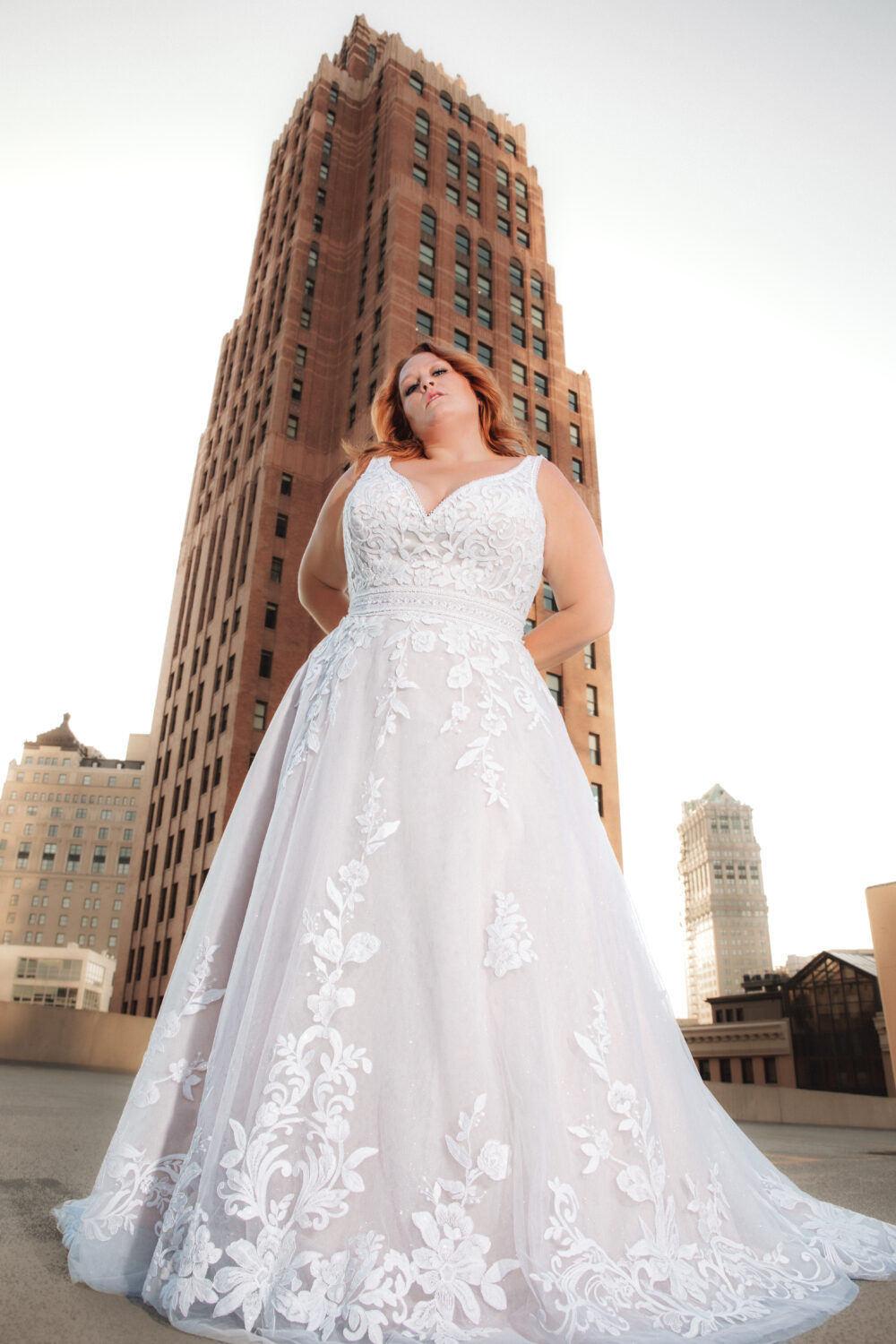 A woman wearing a lace A-line wedding gown stands in front of a tall brick building, posing with her hands behind her back.