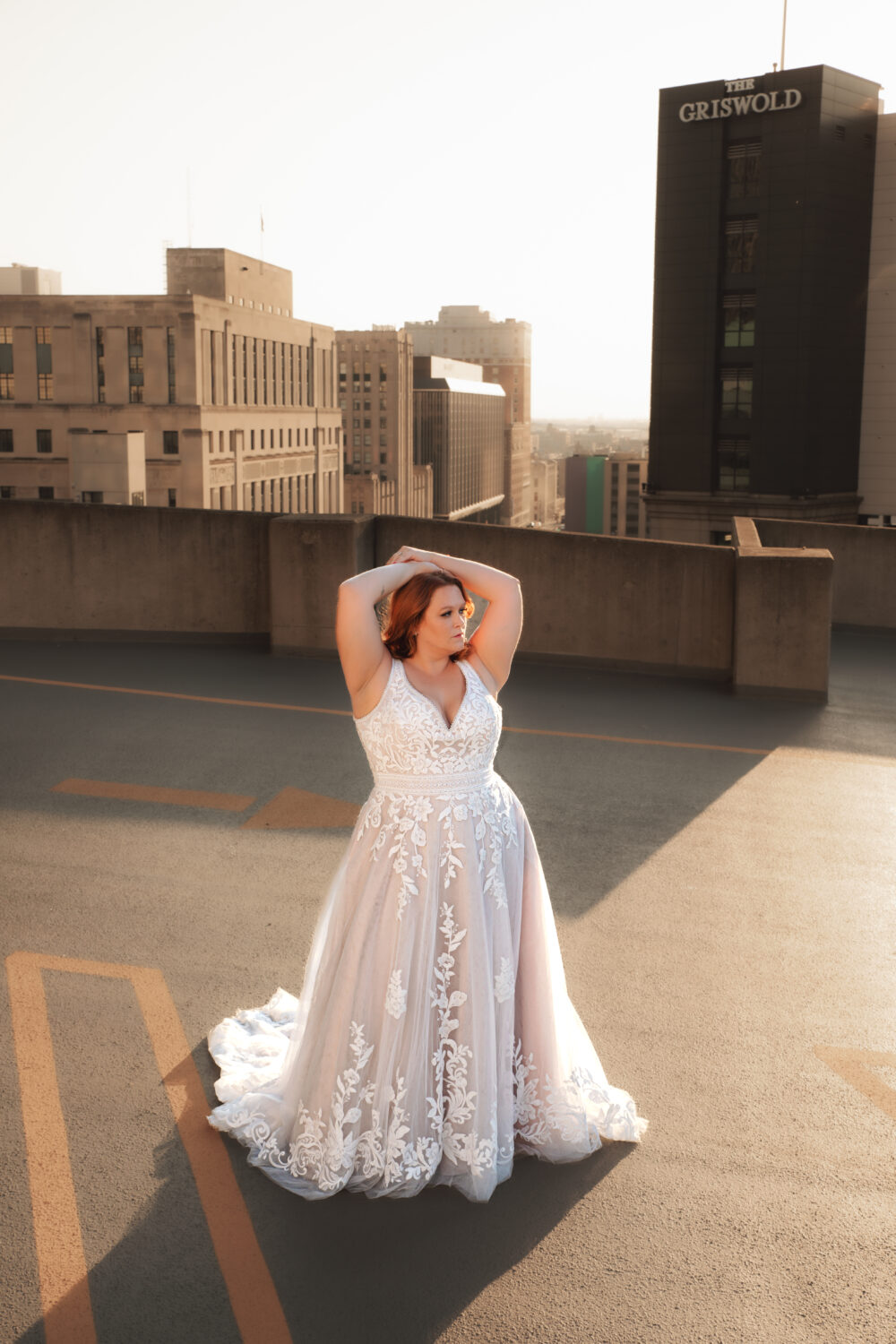 A woman in a lace A-line wedding dress stands on a rooftop with warm golden sunlight behind her, holding her arms raised above her head.